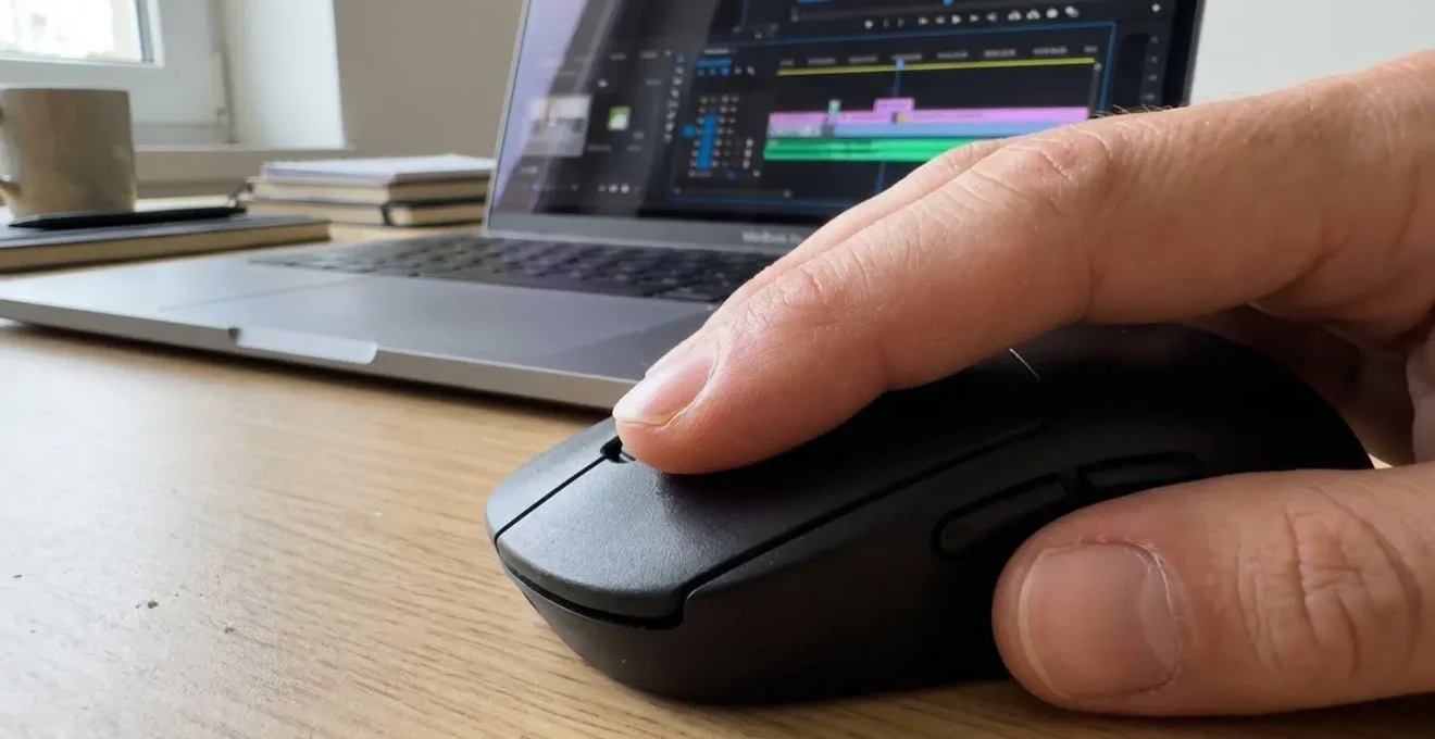 Extreme close-up of a finger clicking a mouse button with a blurred laptop screen showing a video editing timeline in the soft-focus background